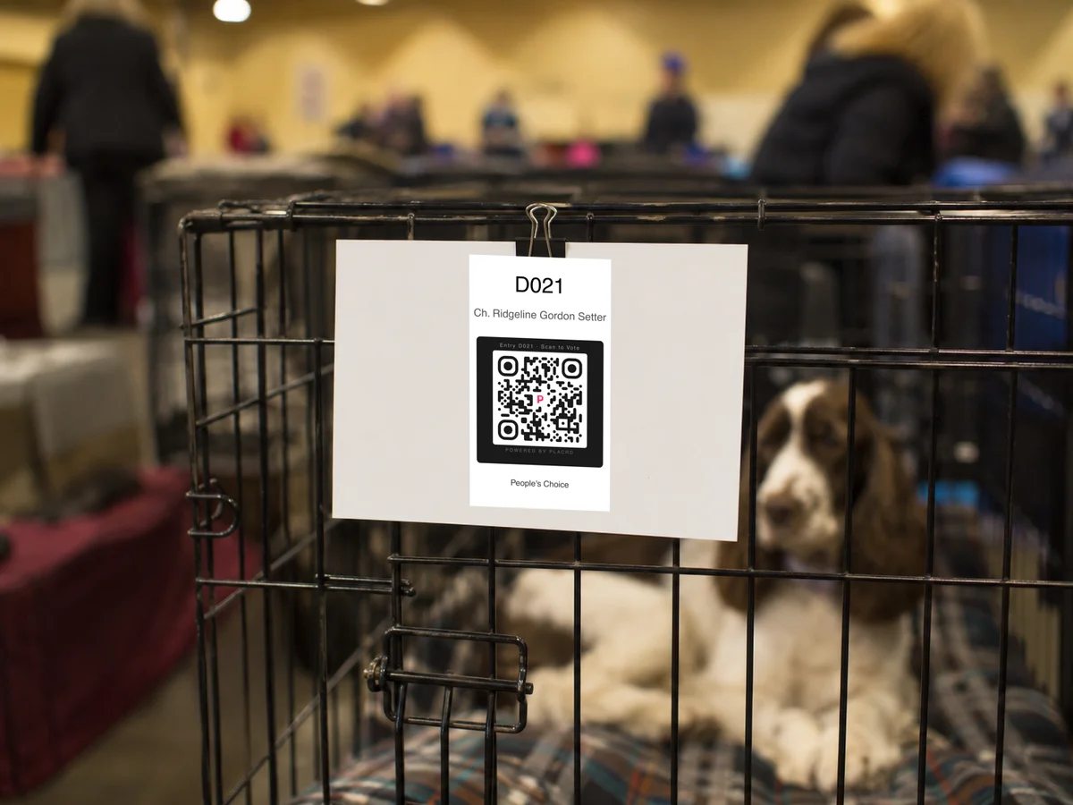 QR voting placard clipped to a dog crate at an indoor dog show